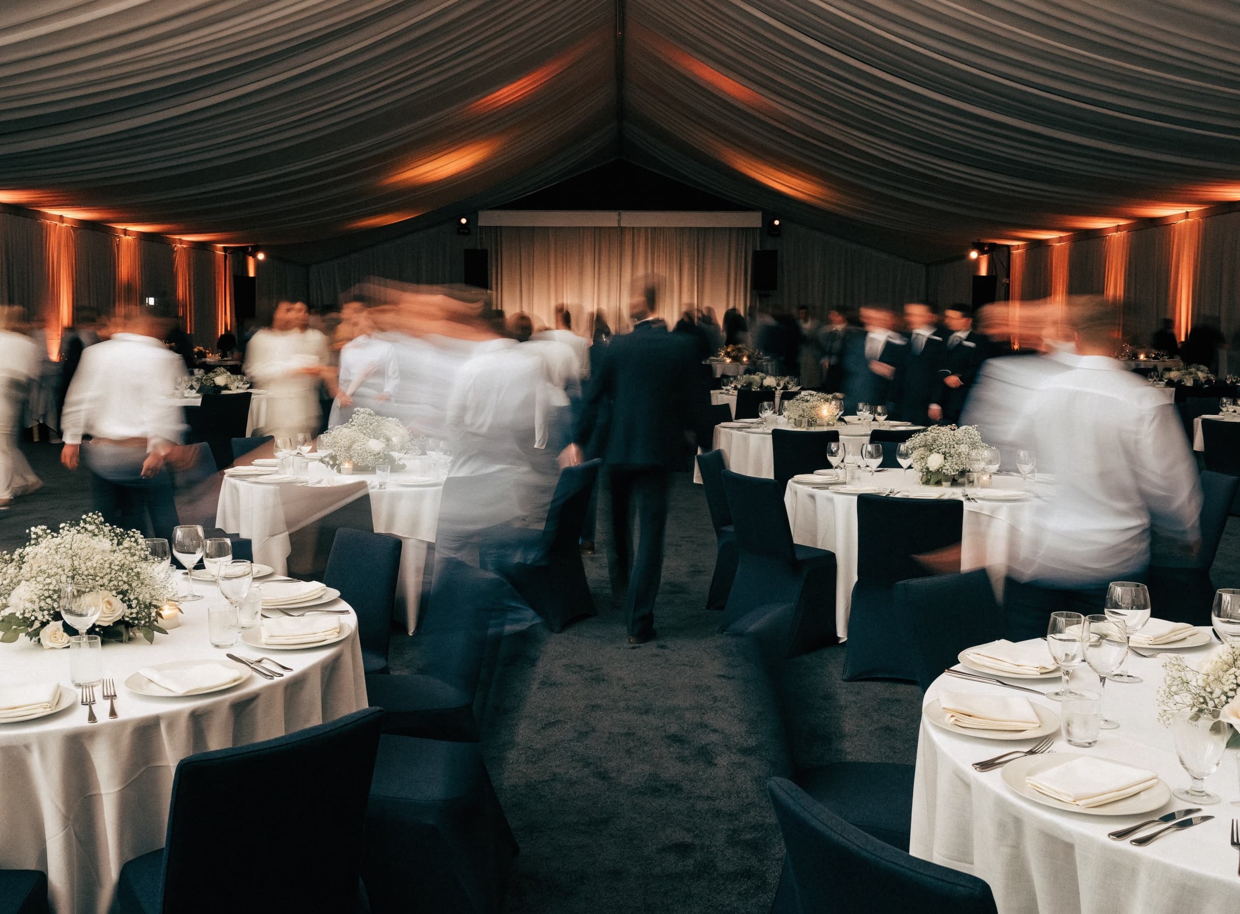A marquee banquet hall during a function, draped ceiling and warm uplighting, guests moving between dressed tables